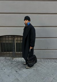 Black overcoat with large lapels, layered over a blue collared shirt. Black bucket hat, black handbag, and grey shoes on a cobblestone street.