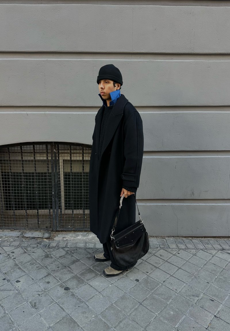 Black overcoat with large lapels, layered over a blue collared shirt. Black bucket hat, black handbag, and grey shoes on a cobblestone street.