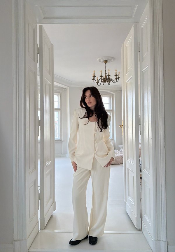 Žena v bílém obleku s černými botami stojící ve dveřích světlé místnosti s lustrem a velkými okny.