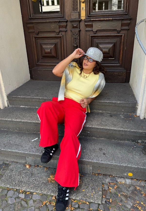 Tenue décontractée comprenant un haut ajusté jaune, un pantalon large rouge avec des rayures blanches et une casquette grise ornée de décorations. Baskets noires.