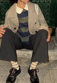 Veste double boutonnage à carreaux marron, polo rayé bleu et vert, pantalon large noir, baskets noires avec des accents rouges et chaussettes à motifs.