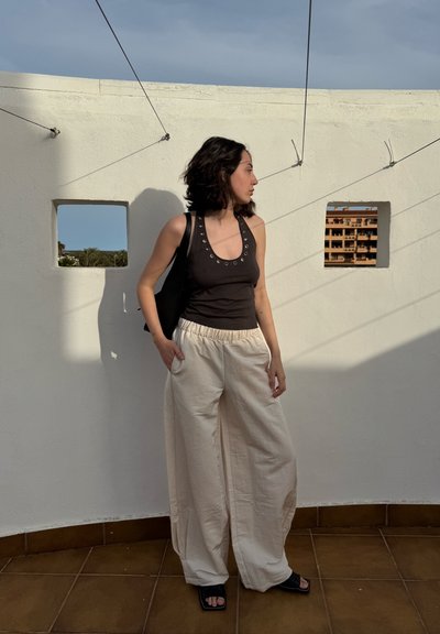 Top negro sin mangas con acentos de ojales metálicos, pantalones de pierna ancha crema y sandalias negras, contra una pared blanca con sombras y una vista parcial de una ventana.