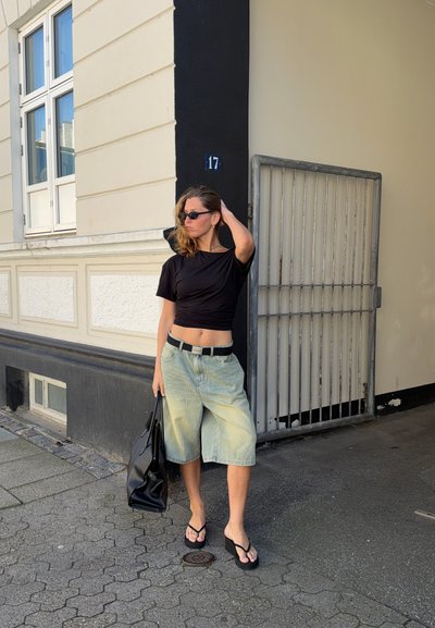 Mujer con top corto negro, pantalones cortos de mezclilla clara hasta la rodilla, sandalias negras y gafas de sol, de pie en la calle sosteniendo una bolsa negra junto a un edificio con el número 17.