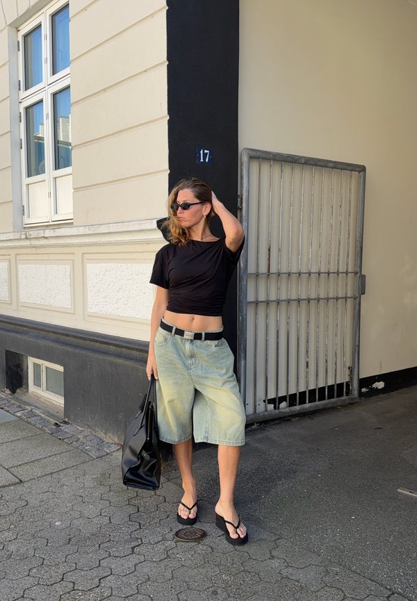 Donna con crop top nero, pantaloncini di jeans chiaro fino al ginocchio, sandali neri e occhiali da sole, in piedi per strada con una borsa nera vicino a un edificio numero 17.