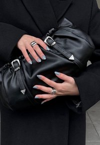 Sac à main en cuir noir avec un design froncé, une quincaillerie argentée et des accents triangulaires. Des mains aux ongles bleu clair tiennent le sac.