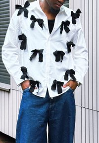 Hvit skjorte med knapper foran, pyntet med svarte stoffsløyfer; brukt over mørkeblå denim bukser med vide ben. Teksturert stoff og lagdelte designdetaljer er synlige.