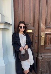 Femme portant des lunettes de soleil, une robe beige et un manteau noir à rayures fines tient un sac à main tissé, appuyée contre une grande porte en bois.