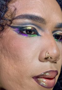 Zbliżenie na twarz prezentującą odważny makijaż oczu z zielonymi, złotymi i fioletowymi akcentami, kolczyk w przegrodzie nosa oraz błyszczące usta w wielotonowych odcieniach.