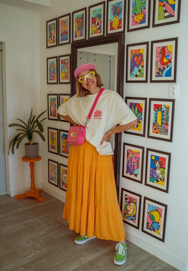 Jeune femme portant un chapeau rose, des lunettes de soleil jaunes, une chemise blanche et une jupe orange, posant en souriant à côté d’un mur d’œuvres d’art encadrées et colorées.