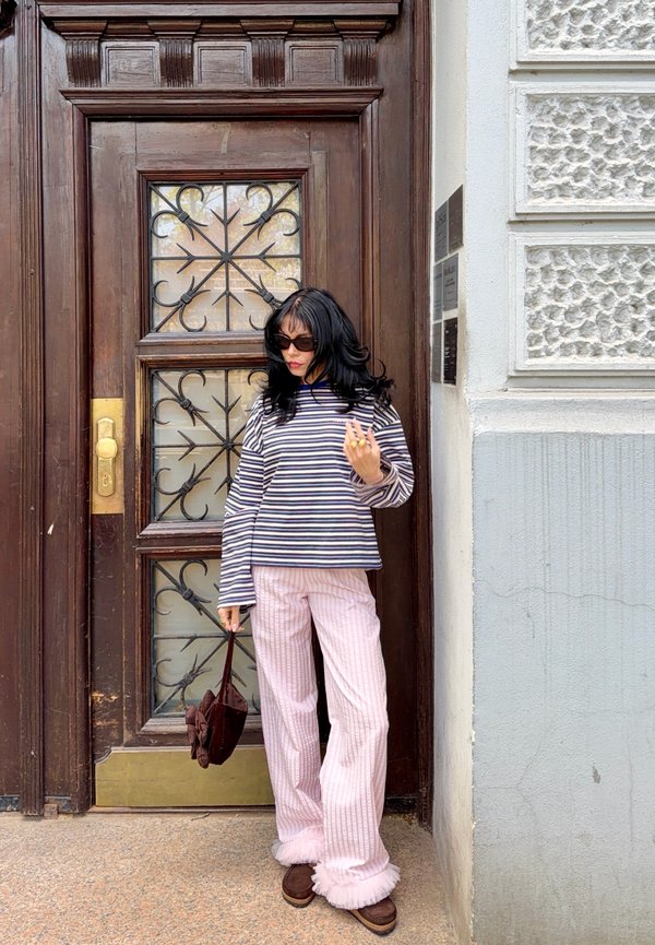 Kobieta o czarnych włosach i w okularach przeciwsłonecznych stoi przy drewnianych drzwiach, ubrana w pasiastą bluzkę z długim rękawem, różowe spodnie i trzyma brązową torbę.