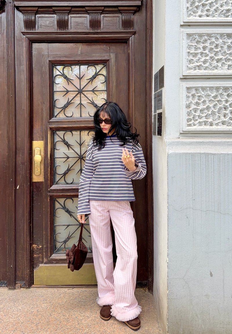 Γυναίκα με μαύρα μαλλιά και γυαλιά ηλίου στέκεται μπροστά σε ξύλινη πόρτα, φοράει ριγέ μακριμάνικη μπλούζα, ροζ παντελόνια και κρατάει καφέ τσάντα.