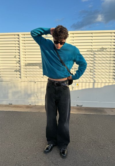 Suéter corto turquesa, pantalones vaqueros negros de cintura alta y zapatos negros brillantes. Contra un fondo claro de tablas con cielo azul.