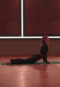 Femeie îmbrăcată în haine negre de antrenament, executând o poziție de yoga cu fața în sus pe un covor verde, într-o cameră cu podea din lemn și panouri de perete cu modele.