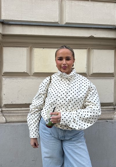Joven con el cabello peinado hacia atrás, vestida con una chaqueta blanca de lunares y vaqueros azul claro, sosteniendo una bebida verde con una pajita negra.