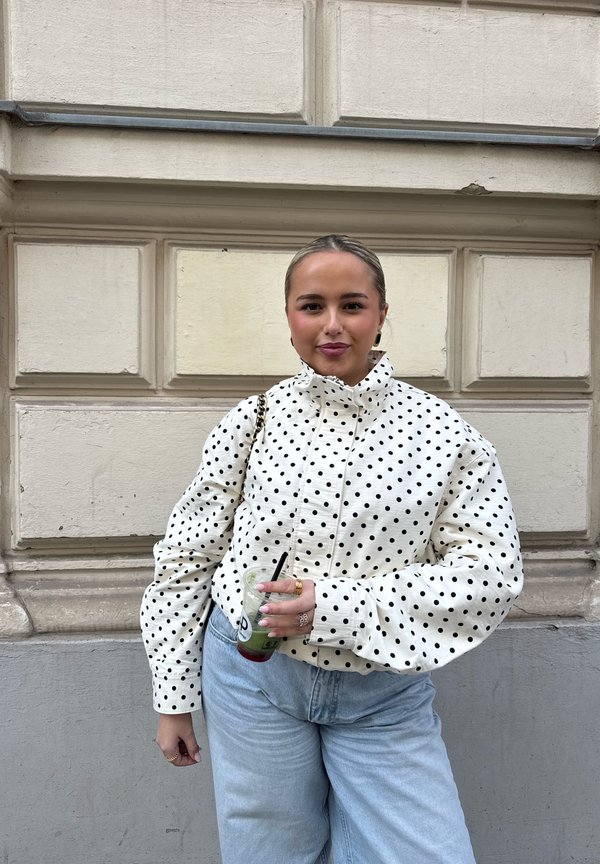 Ung kvinne med bakoverkammet hår iført en hvit jakke med prikker og lyseblå jeans, som holder en grønn drikk med svart sugerør.