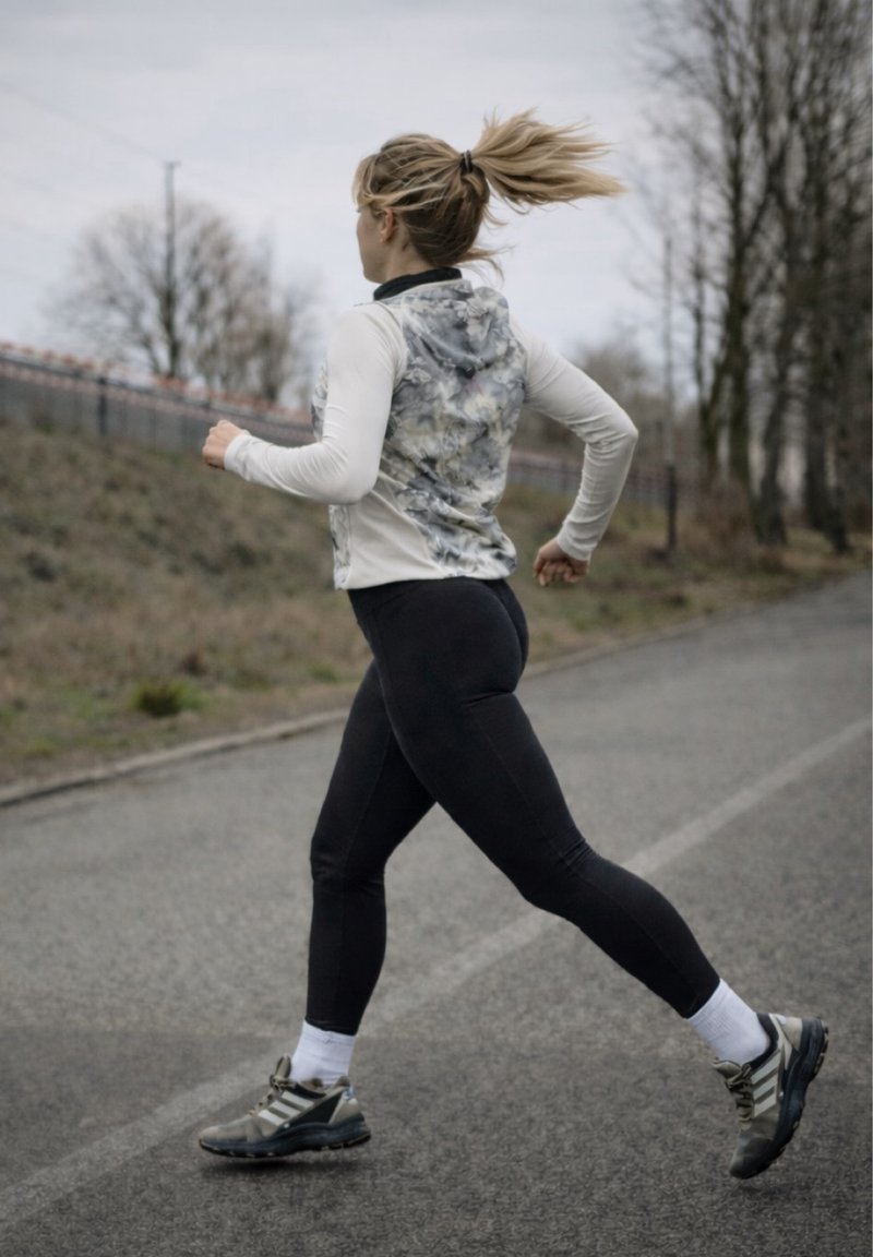 Femme faisant du jogging en plein air sur un chemin pavé, portant des leggings noirs, un gilet à motifs et des baskets, les cheveux attachés en queue de cheval.