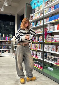 Csíkos, cropped fekete-fehér pulóver, világosszürke, oversized farmernadrág, és sárga platform cipők. Polcok tele könyvekkel és mangákkal.