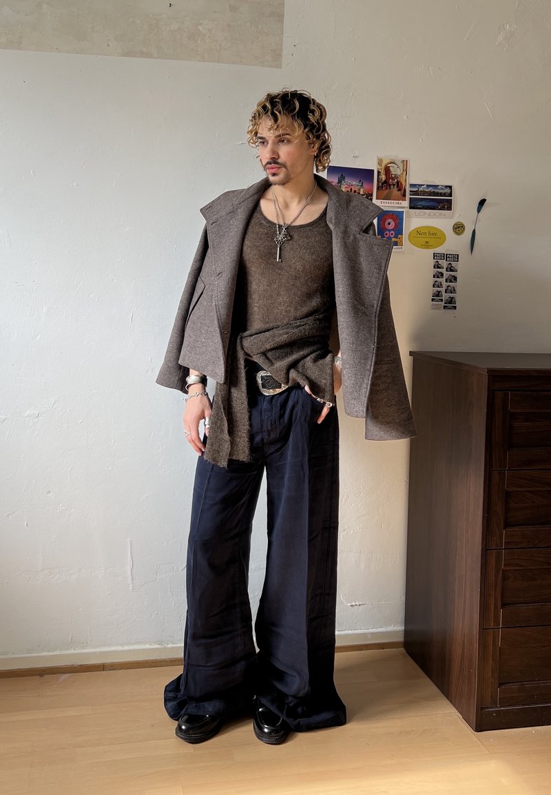 Jeune personne aux cheveux bouclés porte un pull marron et une veste, un pantalon large noir, debout à l'intérieur à côté d'une commode en bois avec des cartes postales sur le mur.