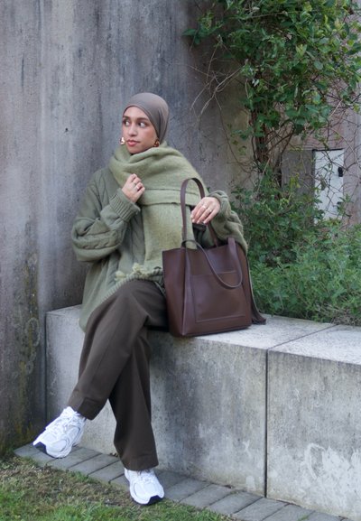 Bolso tote de cuero marrón, suéter de punto verde de gran tamaño, bufanda a juego, pantalones marrones holgados y zapatillas blancas sobre un banco de piedra.