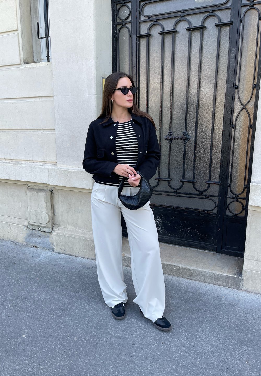 Černá zkrácená bunda přes pruhované tričko, bílé široké kalhoty a černé doplňky. Hladké textury a klasické designové prvky.