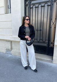 Černá zkrácená bunda přes pruhované tričko, bílé široké kalhoty a černé doplňky. Hladké textury a klasické designové prvky.