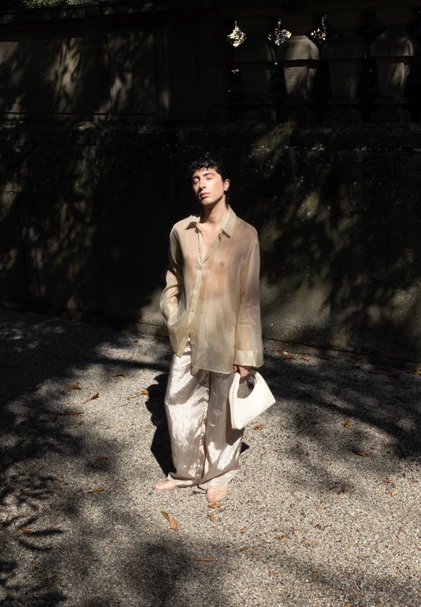 Persona con cabello corto y rizado, vistiendo una camisa beige transparente y pantalones amplios brillantes, se encuentra en un camino de grava, sosteniendo un bolso blanco, con los ojos cerrados bajo la luz del sol.