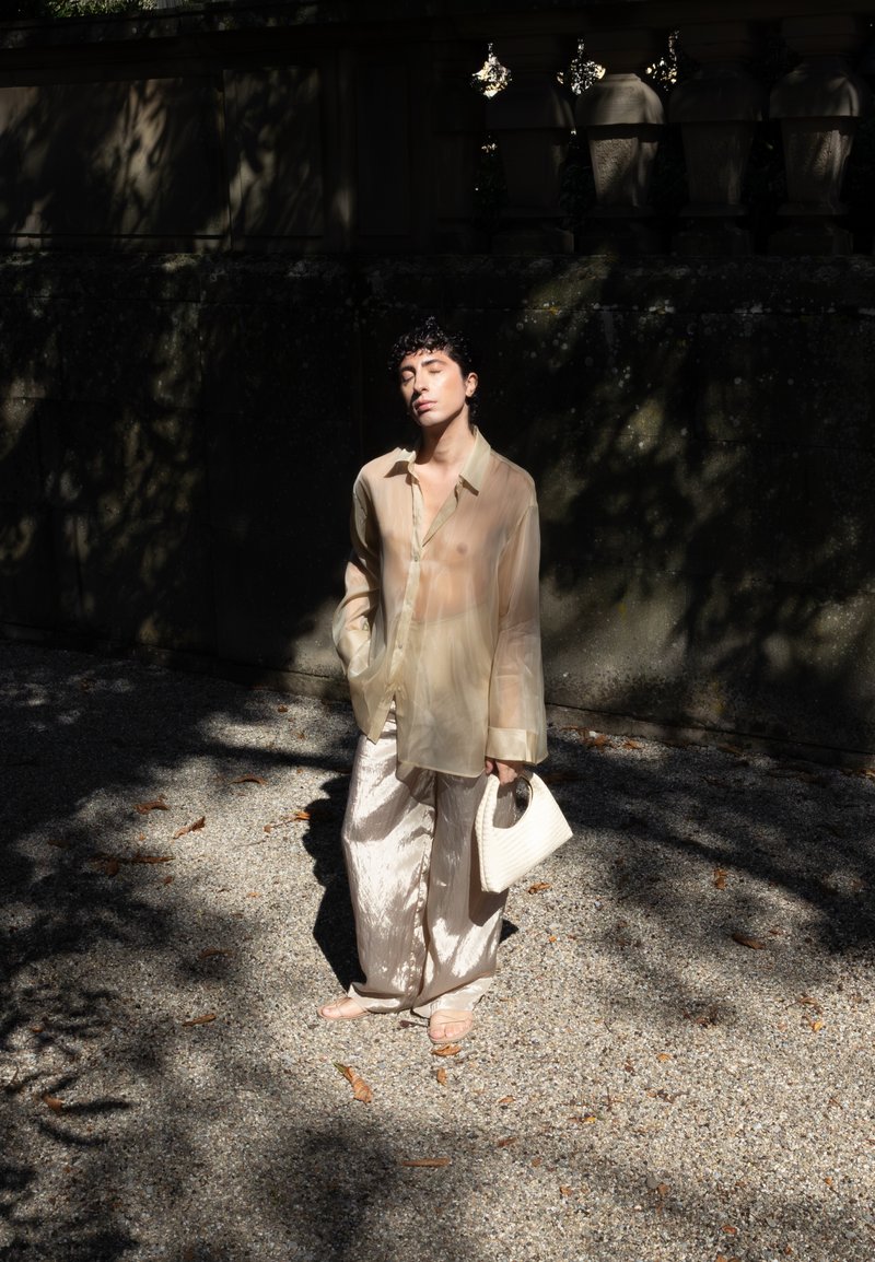 A person with short curly hair is standing on a gravel path, wearing a sheer beige shirt and shiny wide pants. They are holding a white handbag and have their eyes closed, basking in the sunlight.