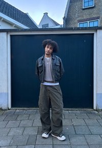 Fekete bőrkabát, szürke kötött pulóver, nagyméretű olívazöld cargo nadrág és fekete sneakers fehér talppal. Sötét háttér előtt áll.