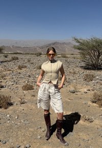 Kvinde iført beige ærmeløs top, hvide shorts, brune støvler og gule solbriller står med hænderne i lommerne i et klippefyldt ørkenlandskab.