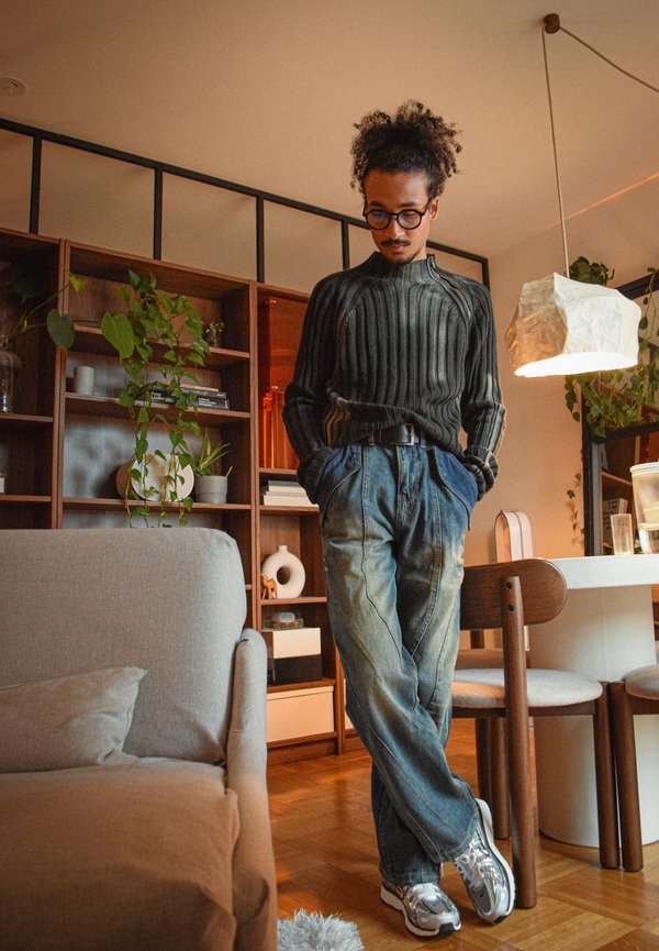 Mann mit lockigem Haar und Brille steht mit den Händen in den Taschen in einem Wohnzimmer mit Holzregalen, Pflanzen und einer hängenden Lampe.