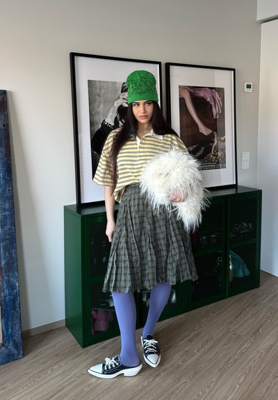 Joven en el interior con sombrero verde, camiseta de rayas, falda de cuadros, medias moradas y zapatos blanco y negro, sosteniendo un bolso blanco y esponjoso.