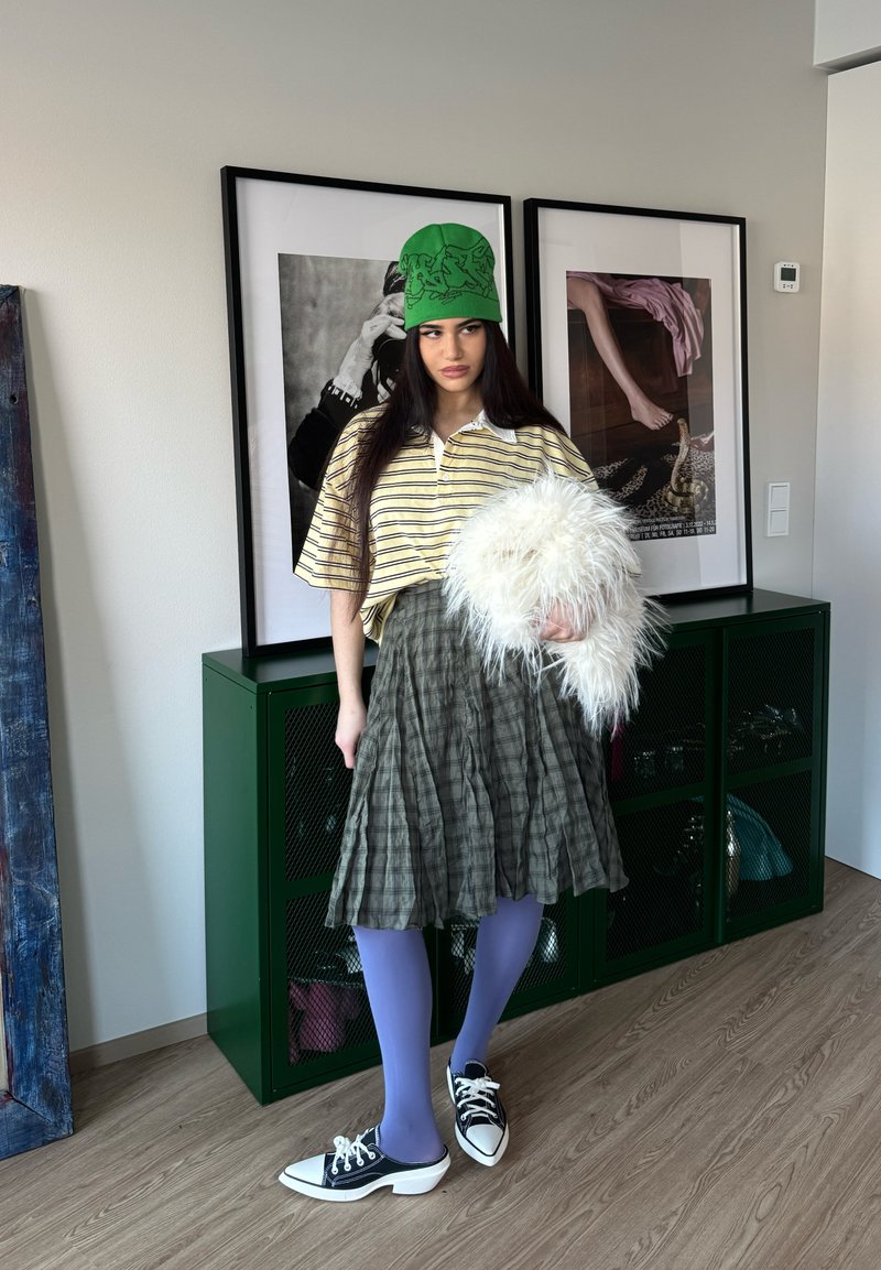 Jeune femme à l'intérieur portant un chapeau vert, une chemise rayée, une jupe à carreaux, des collants violets et des chaussures noir et blanc, tenant un sac blanc et duveteux.