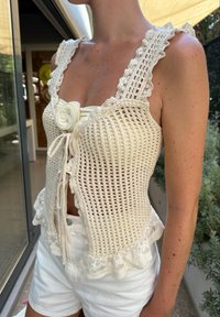 Crochet top in off-white, featuring lace trim, ruffled straps, a floral accent at the centre, and a fitted design. Paired with white shorts.