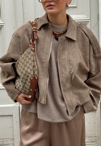 Casaco de camurça castanho com gola, combinado com um suéter taupe e calças de cetim castanhas. Apresenta uma mala de ombro com padrão Gucci.