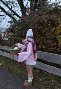 Roze kabelgebreide trui, witte rok met kanten zoom, geruite rode en witte sjaal, beige laarzen met witte sokken, een mand vasthoudend, omgeven door herfstbladeren.