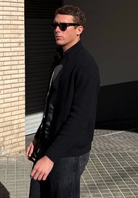 Άνδρας που φοράει μαύρα γυαλιά ηλίου και μαύρο ζακέτα με φερμουάρ στέκεται σε πλακόστρωτο πεζοδρόμιο κοντά σε τοίχο από μπεζ τούβλα κάτω από τον ήλιο.