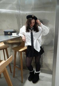 Femme portant un béret noir, une chemise blanche à fines rayures, une cravate noire, une jupe plissée et des bottes doublées de fourrure, s'appuyant sur une chaise près d'une table avec un sac noir et une tasse.