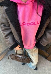 Brun lærjakke med svart pelskant over en rosa hettegenser med teksten "LC DOLLS." Lys blå, slitt jeans i kvart lengde og en brun håndveske.