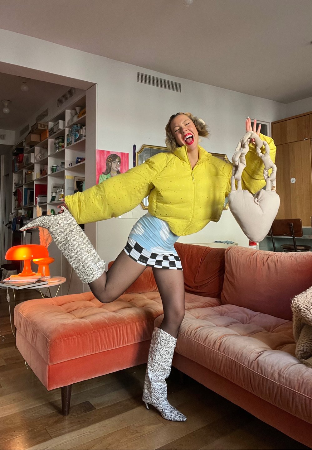 Doudoune jaune oversize avec accents perlés, mini-jupe à carreaux noir et blanc, bottes blanches texturées montant jusqu'aux genoux, sac en forme de cœur.