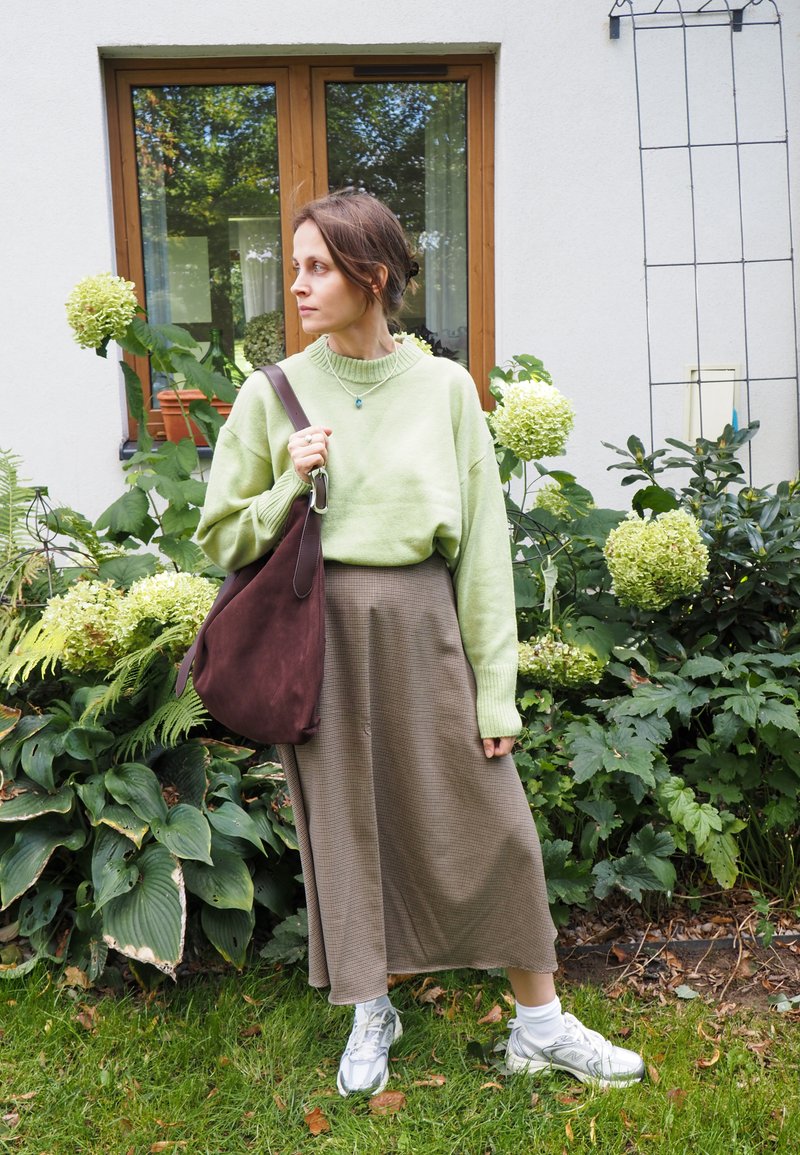 Texturiertes grünes Sweatshirt, brachter Karo-Rock und eine braune Wildledertasche. Silberne Sportschuhe. Umgeben von grünen Blättern und hellen Blumen.