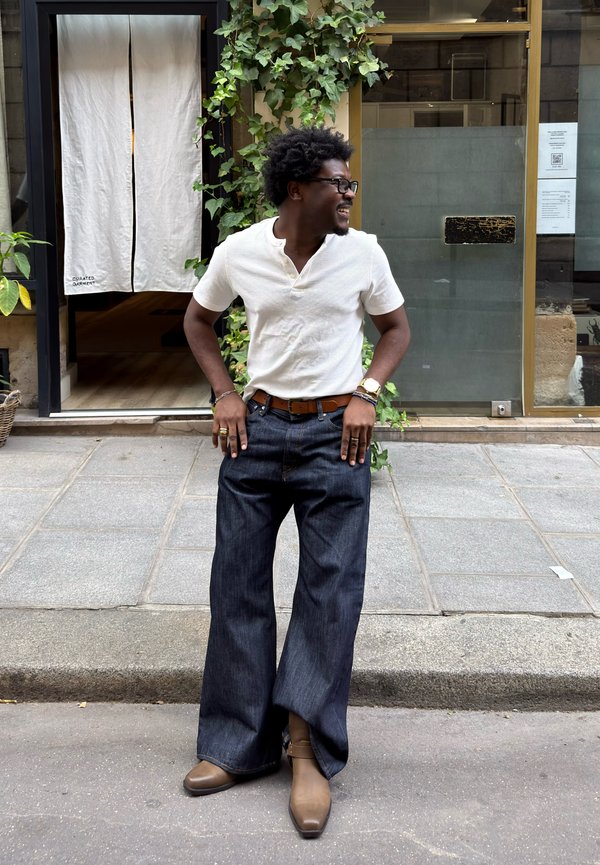 Hombre sonriente con camisa blanca y vaqueros anchos y oscuros de pie en una calle frente a un edificio con hiedra verde y una puerta con cortina blanca.