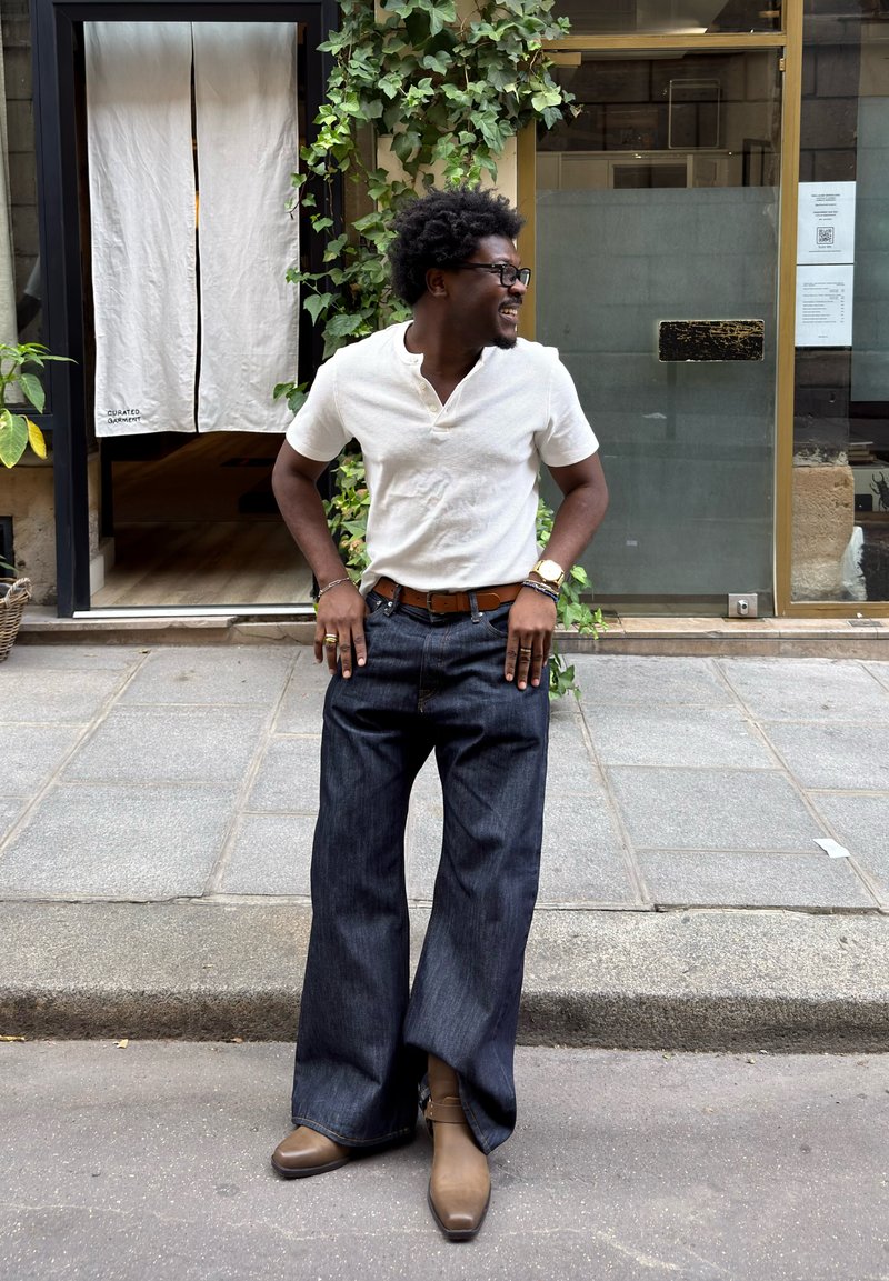 Homme souriant en chemise blanche et large jean foncé debout dans une rue devant un bâtiment avec du lierre vert et une porte avec un rideau blanc.