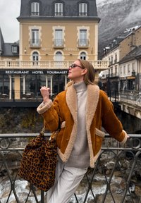 Γυναίκα με γυαλιά ηλίου που φοράει καφέ γούνινο μπουφάν και ανοιχτόχρωμο παντελόνι, στηριγμένη σε μαύρο κάγκελο δίπλα σε ποτάμι, με το ξενοδοχείο Tourmalet στο φόντο.