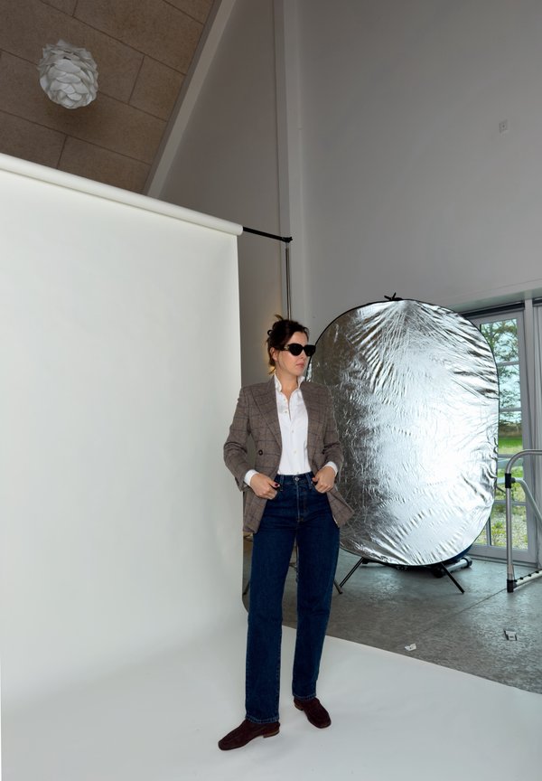 Una mujer con gafas de sol, una blazer de cuadros y vaqueros posa en un estudio fotográfico junto a un gran reflector plateado.