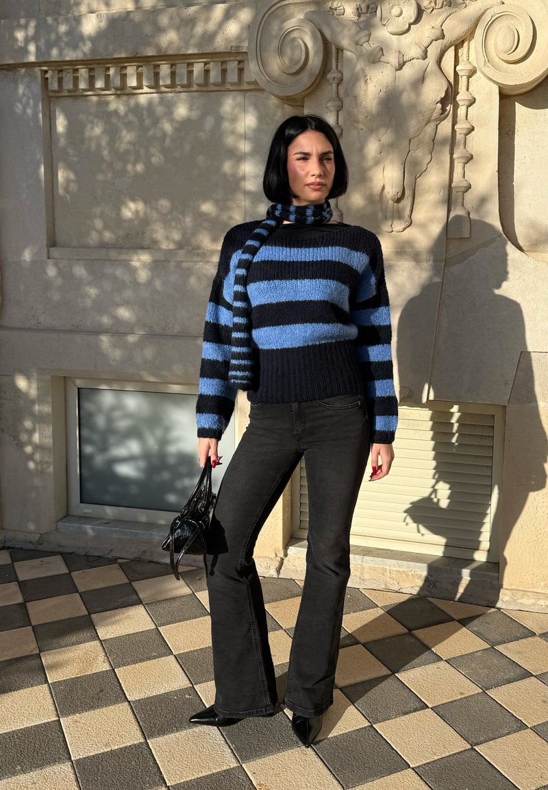 Femme debout sur un pavé à damier, portant un pull et une écharpe rayés noirs et bleus, un pantalon évasé noir, tenant un sac à main noir.