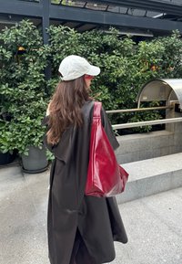 Femme aux longs cheveux bruns portant une casquette blanche et un manteau noir, portant un grand sac cabas en cuir rouge, debout près de buissons verts en plein air.