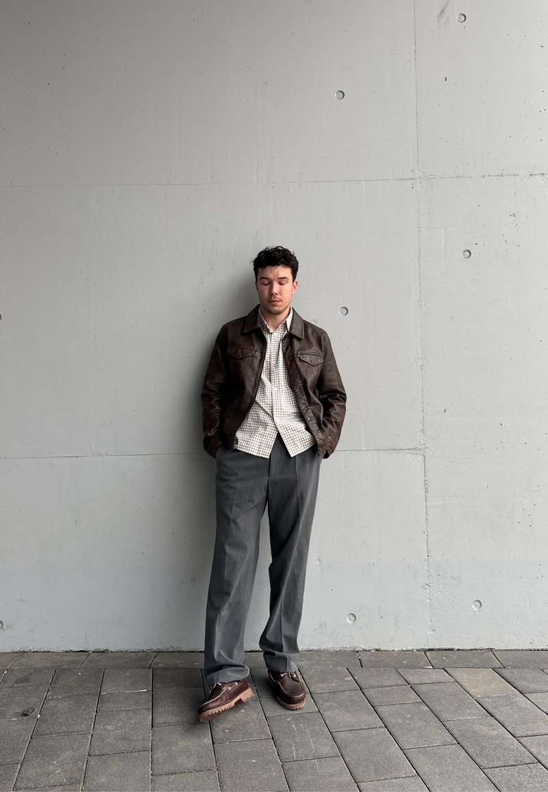 Homem com jaqueta de couro castanho, camisa xadrez, calças cinzentas e sapatos castanhos, de olhos fechados, encostado a uma parede de betão, em um pavimento de azulejos.