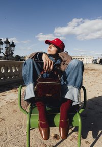 Jeune personne portant une casquette rouge, un manteau à carreaux, des chaussettes rouges et des chaussures marron, assise jambes croisées sur une chaise verte, tenant un sac à main à motifs en plein air.