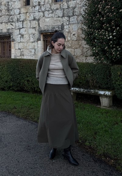 Chaqueta de mezcla de lana en verde oliva, suéter beige de canalé y falda plisada verde oscuro; combinados con botas de tobillo negras; ambiente al aire libre.