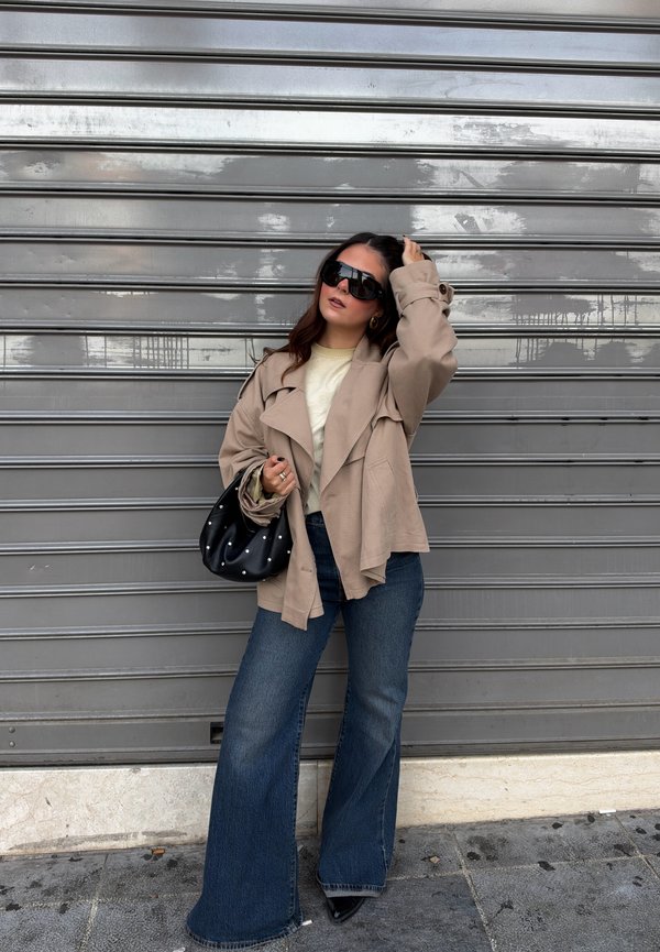 Une femme pose près d’un rideau métallique, portant des lunettes de soleil, une veste beige et un jean à jambes larges, et tenant un sac à main noir clouté.